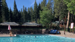 A swimming pool with clear blue water, surrounded by trees and covered seating areas. - A Golden Getaway | Pool, Hot Tub, Tennis, & More (Truckee)