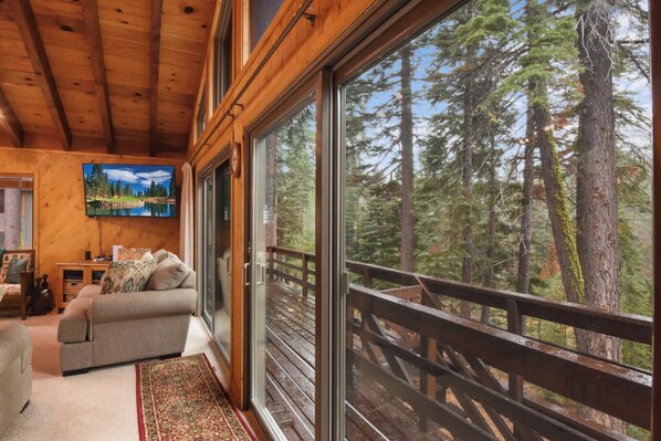 A cozy cabin interior with a large sliding glass door leading to a deck with a wooden railing, offering a view of tall trees.