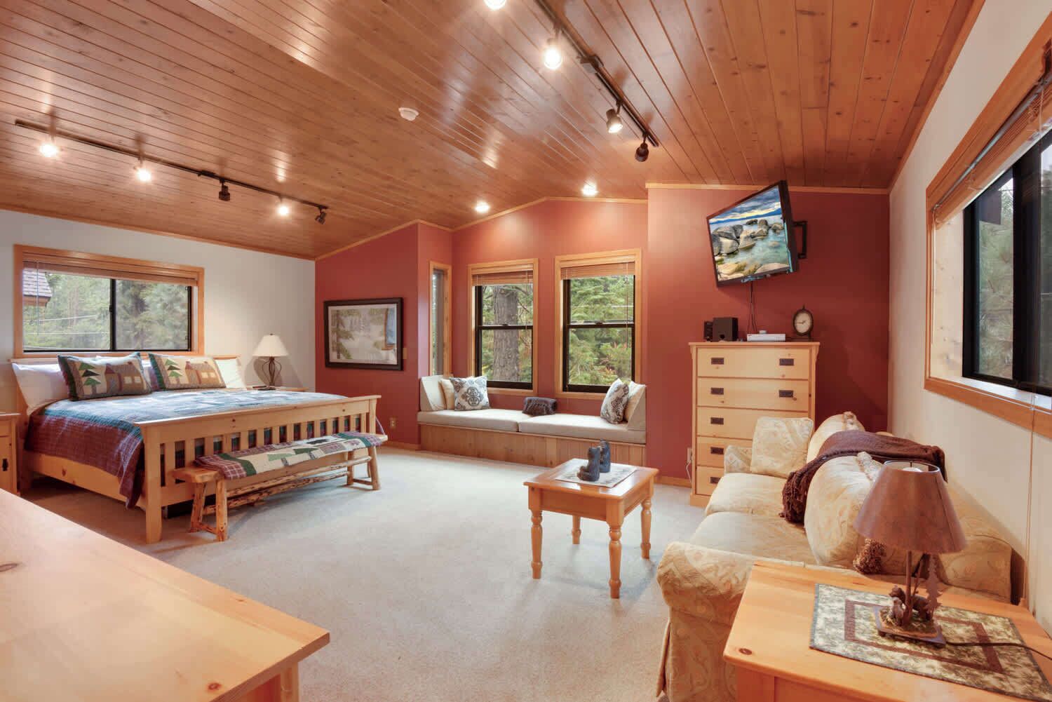 Une chambre confortable avec un plafond en bois, un grand lit, un coin salon avec un banc, une commode et un téléviseur fixé au mur.