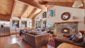 A spacious living room with a stone fireplace, leather sofas, wooden beams, and a large window overlooking a forested area. - Northstar Treehouse – Chic 4BR House | Forest view in Northstar Ski Resort (Truckee)