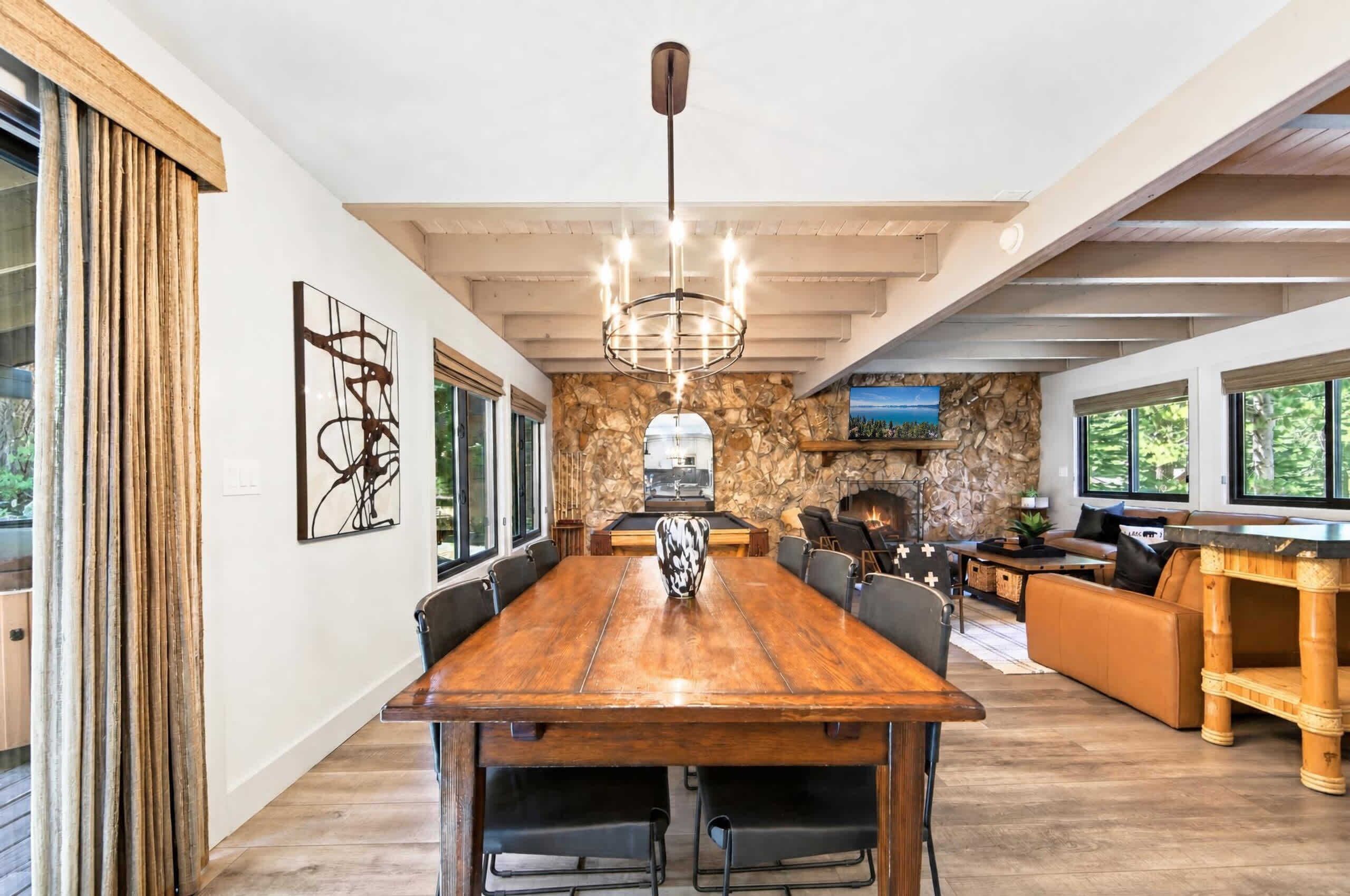A spacious living area with a large wooden dining table, comfortable seating, a stone fireplace, and a modern kitchen in the background.
