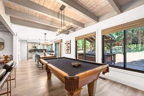 Un espacio interior amplio con mesa de billar, piso de madera y grandes ventanales con vistas al exterior.