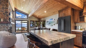 A modern kitchen with a large island, stone wall, wooden ceiling, and large windows overlooking a scenic view. - Shoreline Vista | 180° Lake Views | Pet-Friendly | 2 Master Suites (Carnelian Bay)