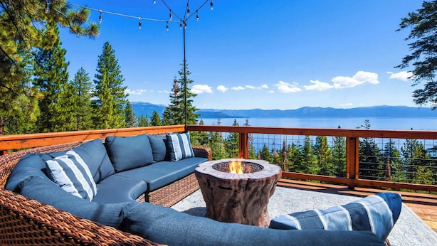 A wicker sofa with blue cushions, a round wooden table with a fire pit, and a view of a lake and mountains. - Shoreline Vista | 180° Lake Views | Pet-Friendly | 2 Master Suites (Carnelian Bay)