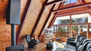 A cozy cabin interior with a fireplace, leather sofa, and a large window overlooking a deck.