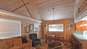 A cozy cabin interior with a wooden ceiling, a dining area, a living room with a sofa and armchairs, and a kitchen area with a bar. - Woodland Chalet | 2 mi from Beach Access! (Tahoma)