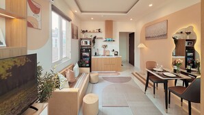 Un salon moderne avec un téléviseur, un canapé et une table basse, un coin repas avec des chaises et une table, et une cuisine équipée d’appareils électroménagers et dotée d’une fenêtre.
