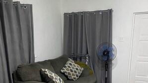 Una habitación con cama, sofá, ventilador de pie y cortinas grises.