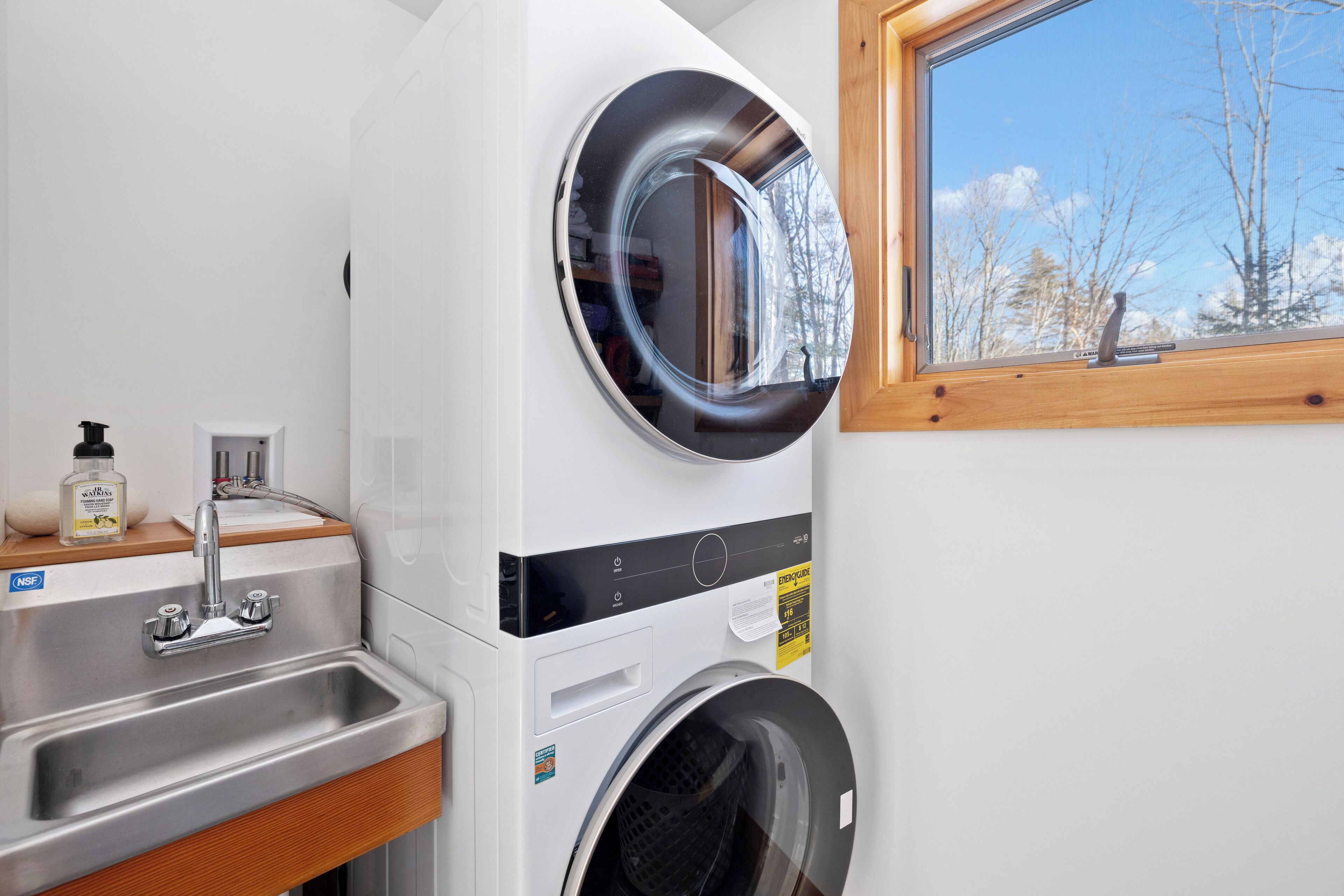 Een kleine wasruimte met een wasmachine en droger, een wastafel en een raam met uitzicht op bomen.