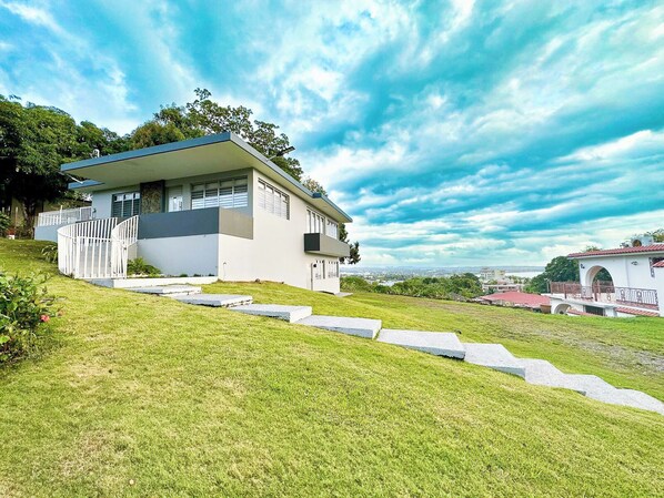 Una casa moderna con techo plano, fachada blanca y un pequeño balcón. Un césped delantero con un sendero de piedra que lleva a la entrada.