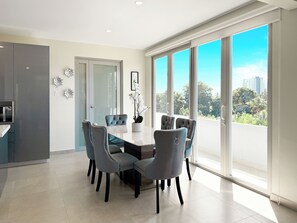 Un moderno comedor con una gran mesa y seis sillas, una cocina con horno empotrado y ventanas amplias con vistas a la ciudad.