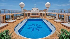 A cruise ship deck with a central pool, sun loungers, and a bar area.