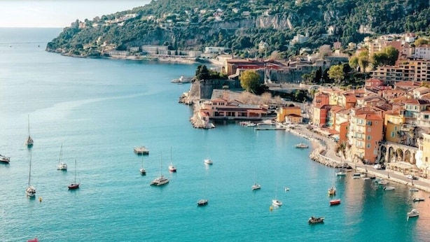 A coastal town with a marina filled with boats, buildings with Mediterranean architecture, and a hilly backdrop.