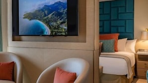 A hotel room with a large flat-screen TV mounted on a wooden panel, a bed with a teal headboard, two armchairs with cushions, and a nightstand with a lamp.