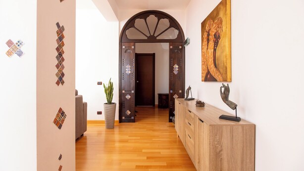 A hallway with wooden flooring, a decorative archway, a potted plant, and a wooden cabinet with a sculpture on top. - LuminArt (Gibellina)