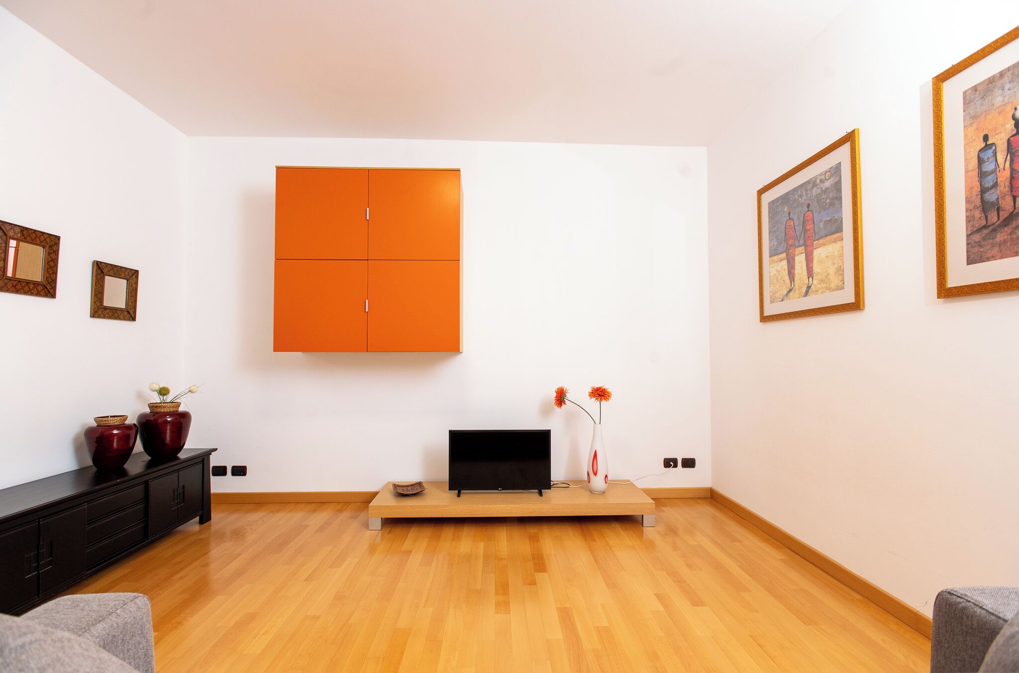 Un salon moderne avec un plancher en bois, un téléviseur noir posé sur un meuble en bois, un buffet orange et un vase rempli de fleurs.