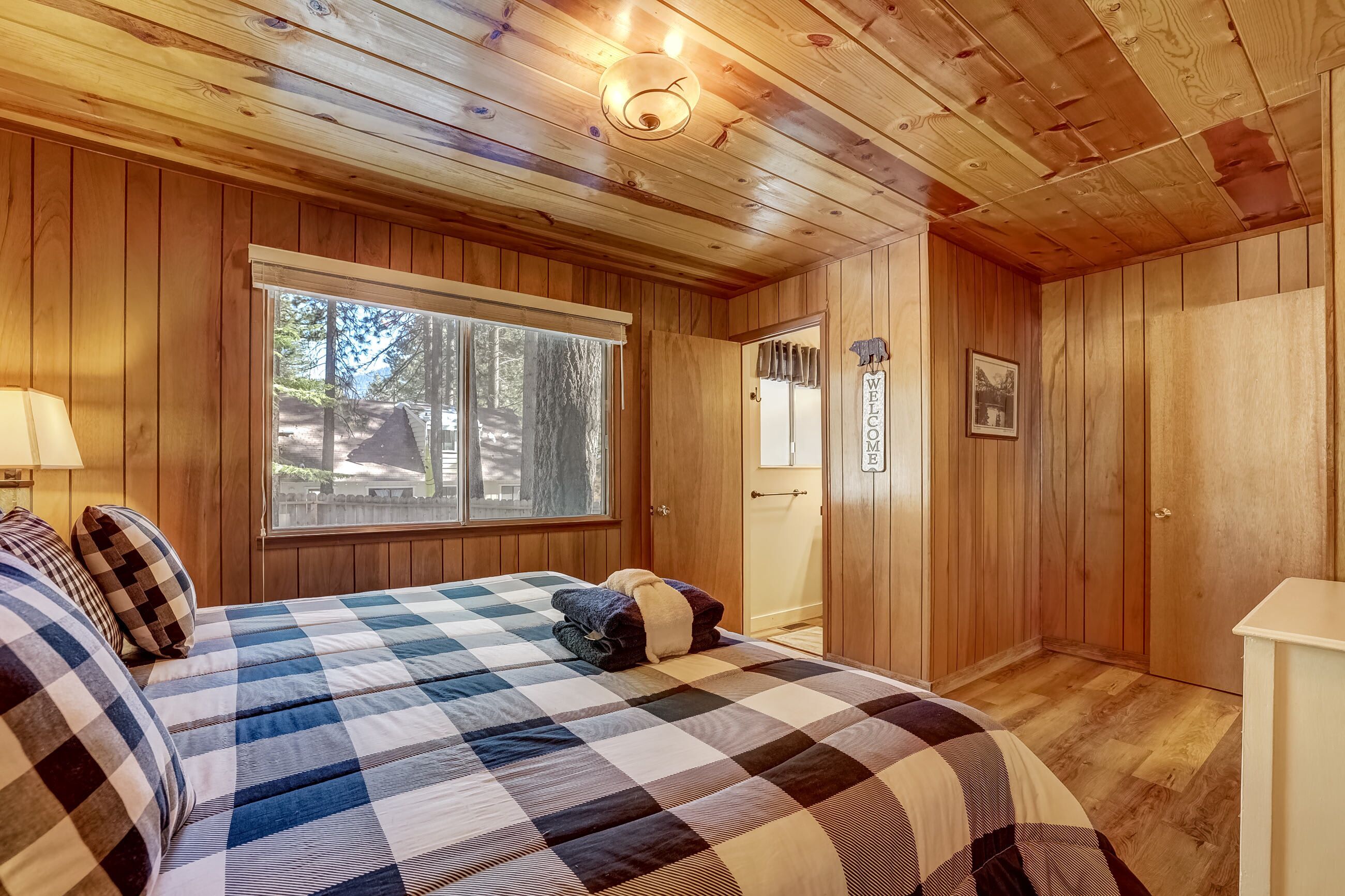 A wooden cabin room with a checkered bed, a window, a door, and a welcome sign.