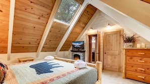 Une chambre confortable située sous les toits, avec un plafond en pente, un grand lit recouvert d’une courtepointe, une cheminée, des meubles en bois et une vue sur l’extérieur grâce à une lucarne.