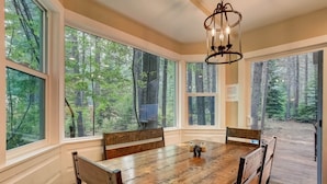 Un espace repas comprenant une grande table et des chaises en bois, de grandes fenêtres donnant sur la forêt et un lustre.