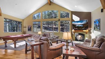 A spacious living room with a pool table, leather sofas, a fireplace, and large windows offering a forest view.
