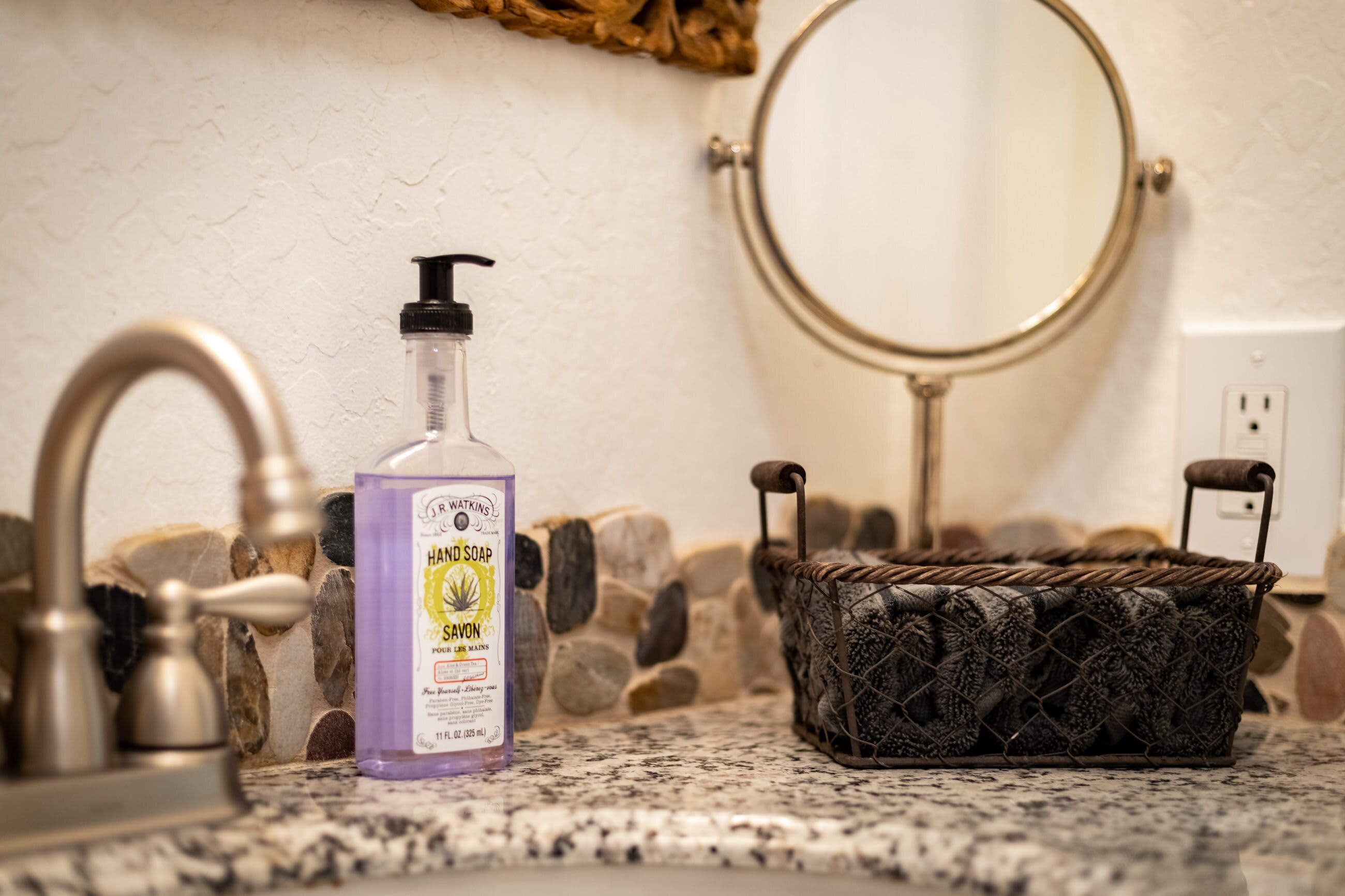 A bathroom with a granite countertop, a soap dispenser, a round mirror, and a stone wall.