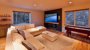 A modern living room with a sectional sofa, a flat-screen TV, a piano, and large windows with a view of trees. - The Elevator House – Sleeps 10! (Stateline)