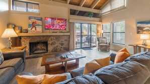 A spacious living room with a stone fireplace, a television mounted above it, and a large window overlooking a deck. - Lakeland Sandy Beach Retreat | Private Escape (South Lake Tahoe)