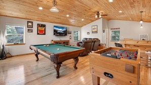 A spacious room with a pool table, a foosball table, a television, and a ceiling fan.