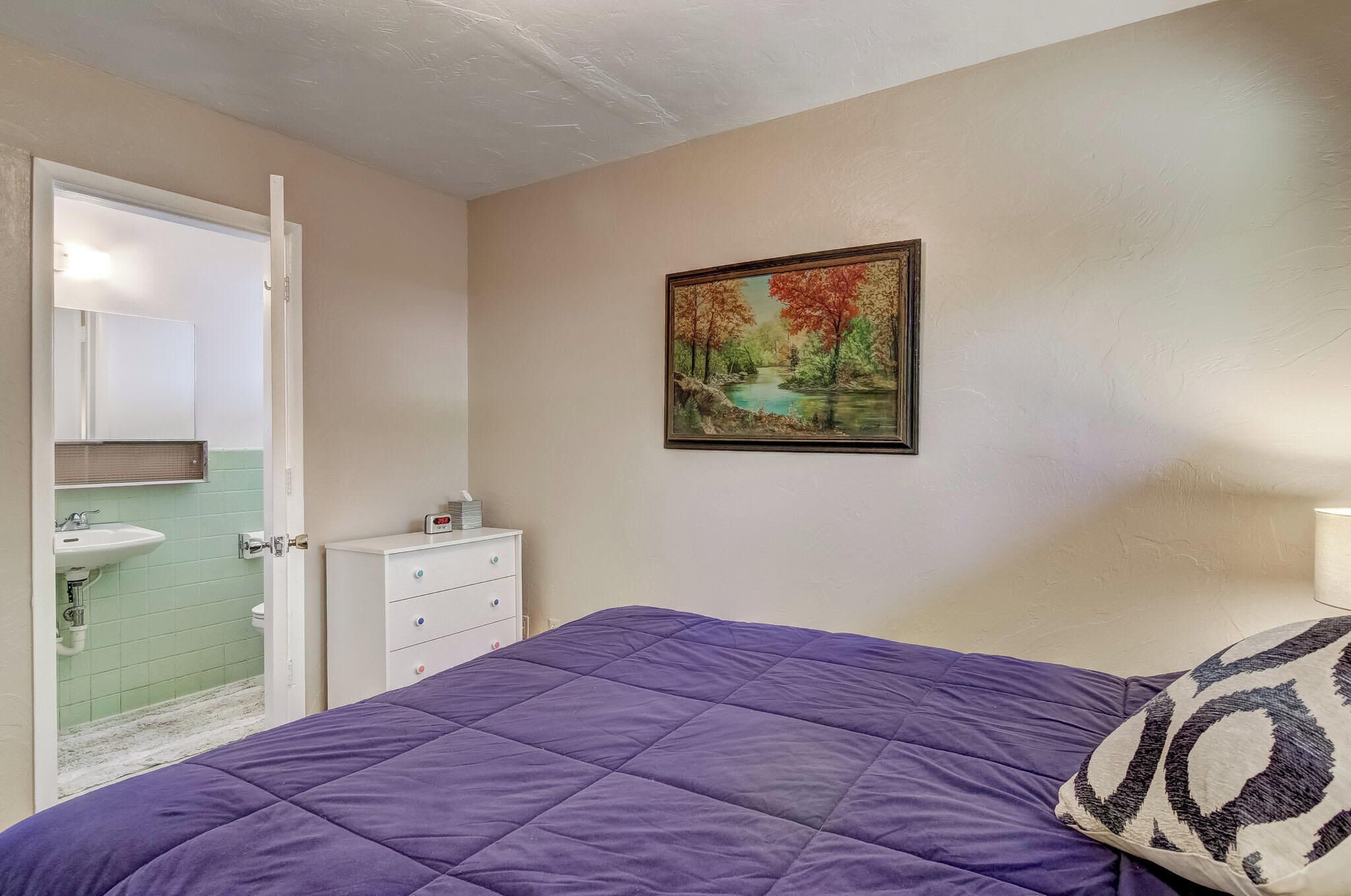 Une chambre à coucher avec une couvre-lit violette, une commode blanche, un tableau représentant un paysage, et une salle de bain avec un lavabo vert et des carreaux blancs.