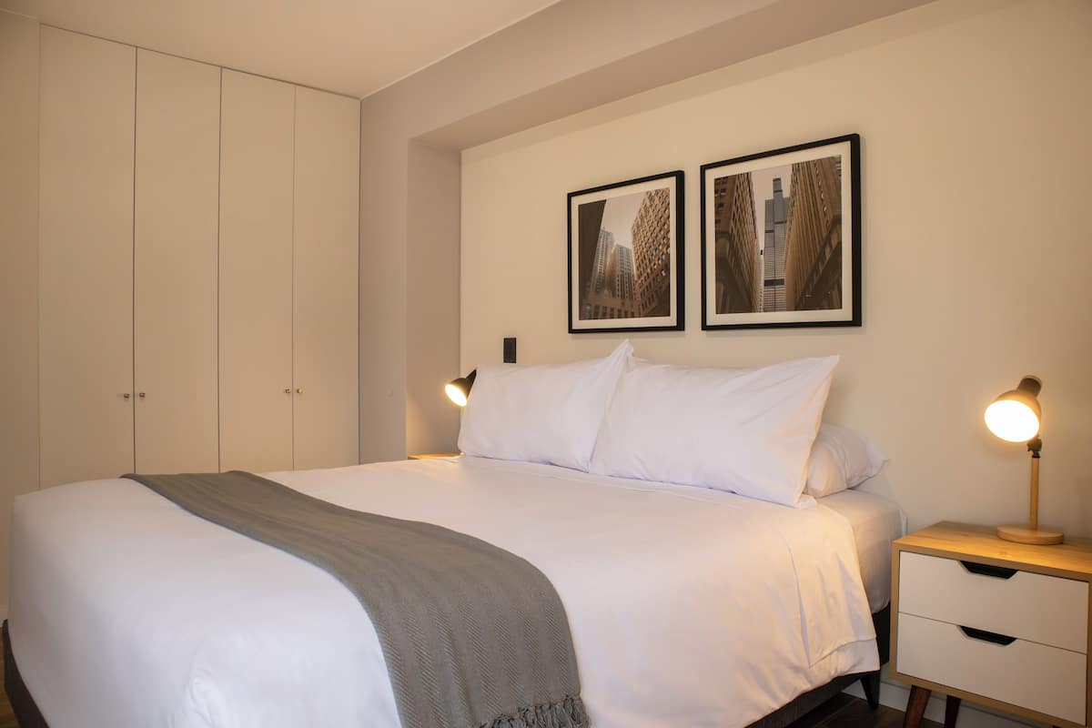 Un lit bien fait, recouvert d’un plaid gris, une table de chevet avec une lampe, et deux tableaux encadrés accrochés au mur.