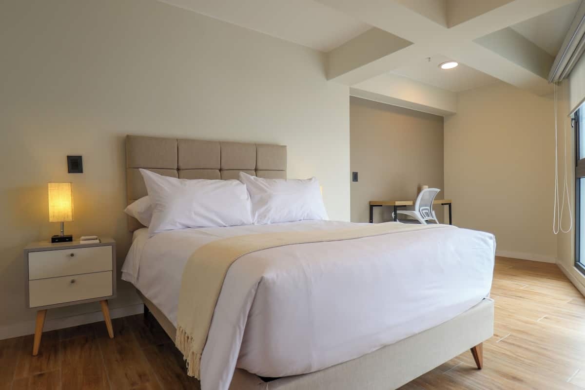 Une chambre à coucher moderne avec un grand lit, une table de chevet avec une lampe et une vue sur l’extérieur par une fenêtre.