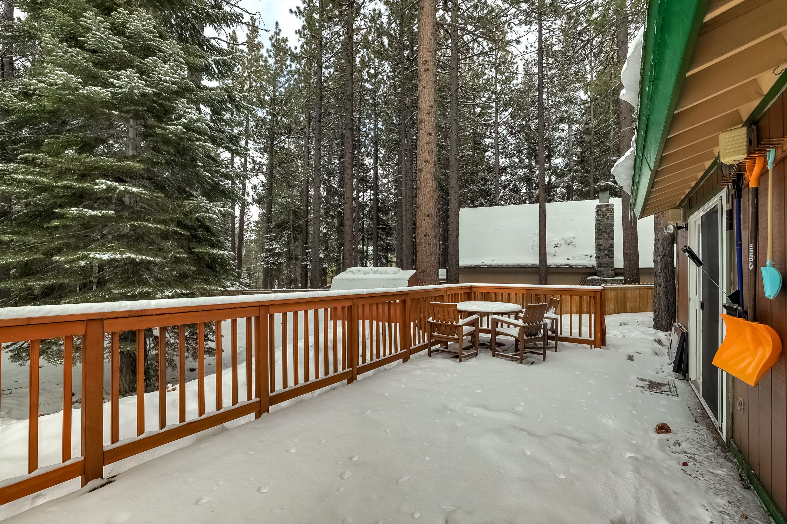 A wooden deck with a table and chairs, surrounded by snow-covered ground and tall trees.