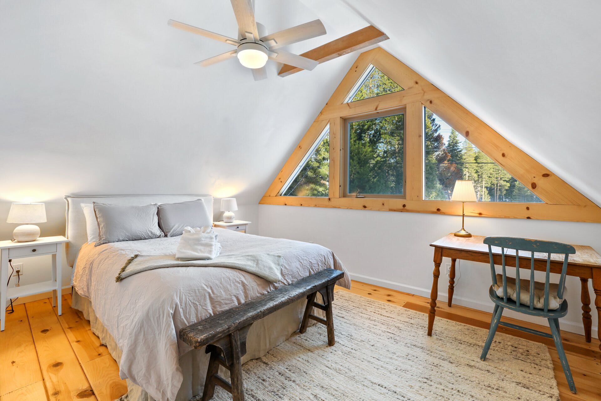 Un dormitorio con techo inclinado, una cama, un escritorio, una silla y una ventana con vista a árboles.