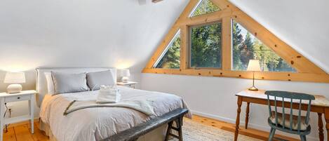 Un dormitorio con techo inclinado, una cama, un escritorio, una silla y una ventana con vista a árboles.