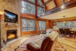 Una sala de estar acogedora con chimenea de piedra, ventanas grandes, un sofá y un comedor.