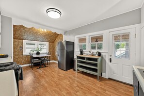 A kitchen with a wooden floor, a refrigerator, a dining table with chairs, and a window with a view of greenery.