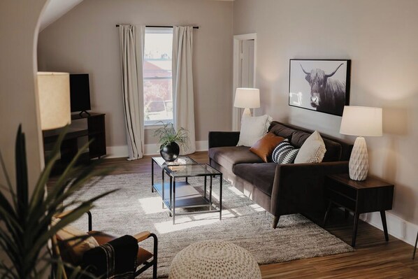 A living room with a sofa, two side tables, a coffee table, a TV, and a window with curtains.