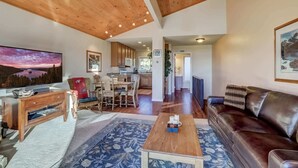 A spacious living room with a wooden ceiling, a large flat-screen TV, a brown leather sofa, a wooden coffee table, and a blue patterned rug.
