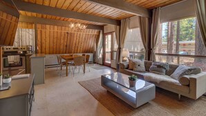 Een modern interieur met een houten plafond, een keuken, een zithoek met bank en salontafel, en grote ramen met uitzicht op bomen en gebouwen.