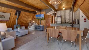Een ruime woonkamer met houten plafond, een open haard, een televisie en een eettafel met stoelen.