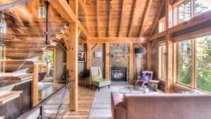 A spacious cabin interior with a stone fireplace, wooden beams, and large windows offering a view of the outdoors.