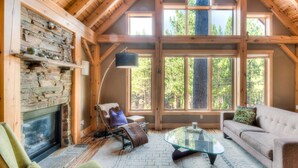 A cozy cabin interior with a stone fireplace, wooden beams, large windows, and a comfortable seating area.