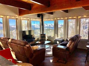 Un salon chaleureux avec des canapés en cuir, un téléviseur à écran plat et un poêle à bois. De grandes fenêtres offrent une vue sur les montagnes.