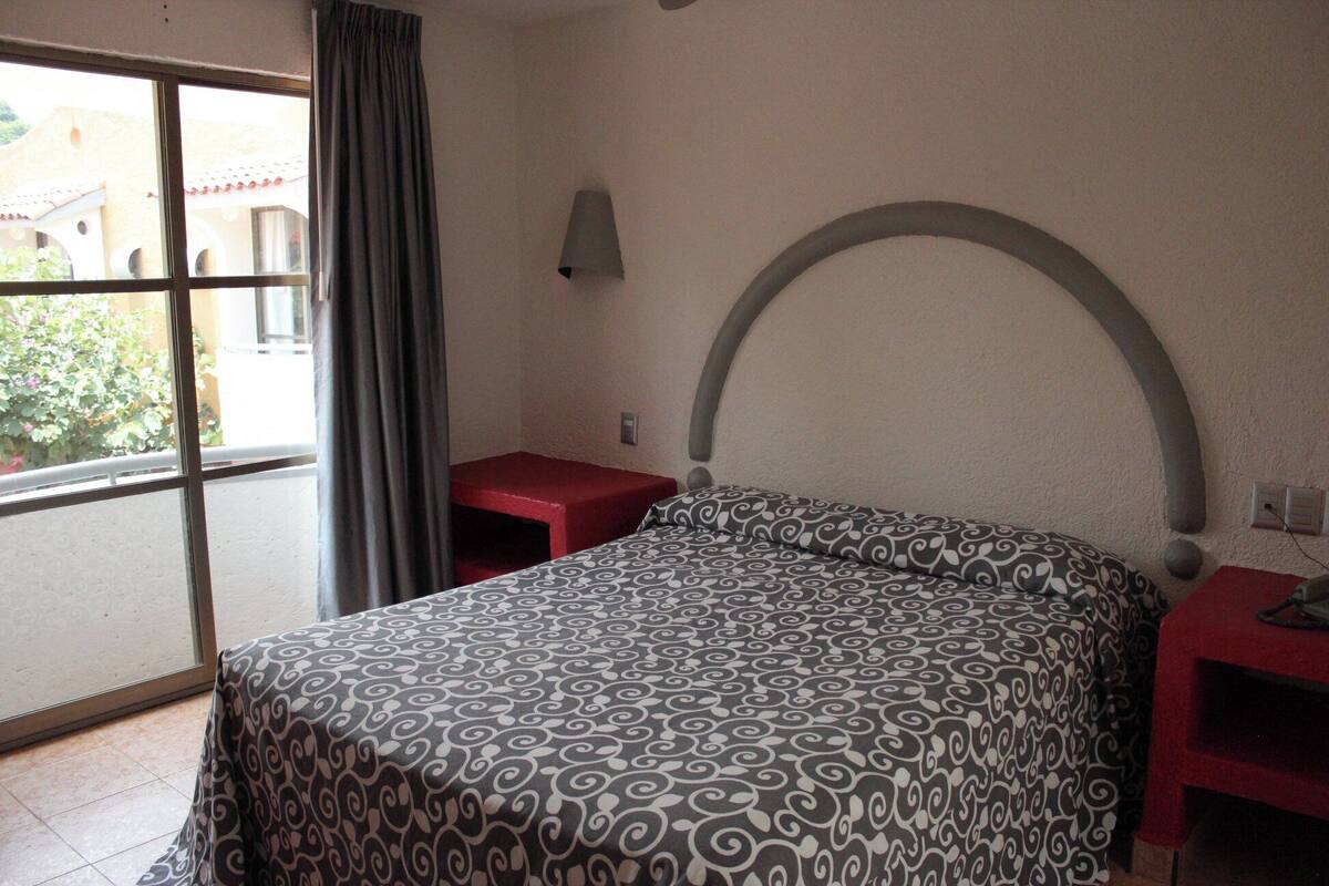 A hotel room with a bed, a small red table, a wall-mounted lamp, and a window with a view of buildings and greenery.