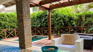 A poolside area with a stone pillar, a glass table with a bowl, white wicker furniture, and lush greenery in the background. - Luxury 2-Bedroom Apartment with Private Mini Pool & Backyard in Las Terrenas (El Limón)