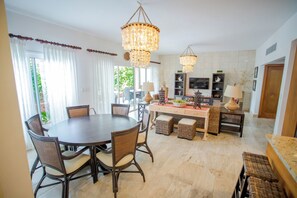 A dining area with a round table and chairs, a kitchen with a TV and shelves, and a large window with curtains.