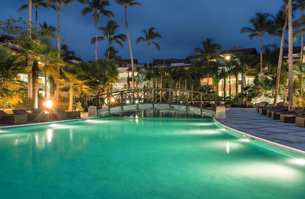 A swimming pool with a wooden bridge, surrounded by palm trees and hotel buildings.