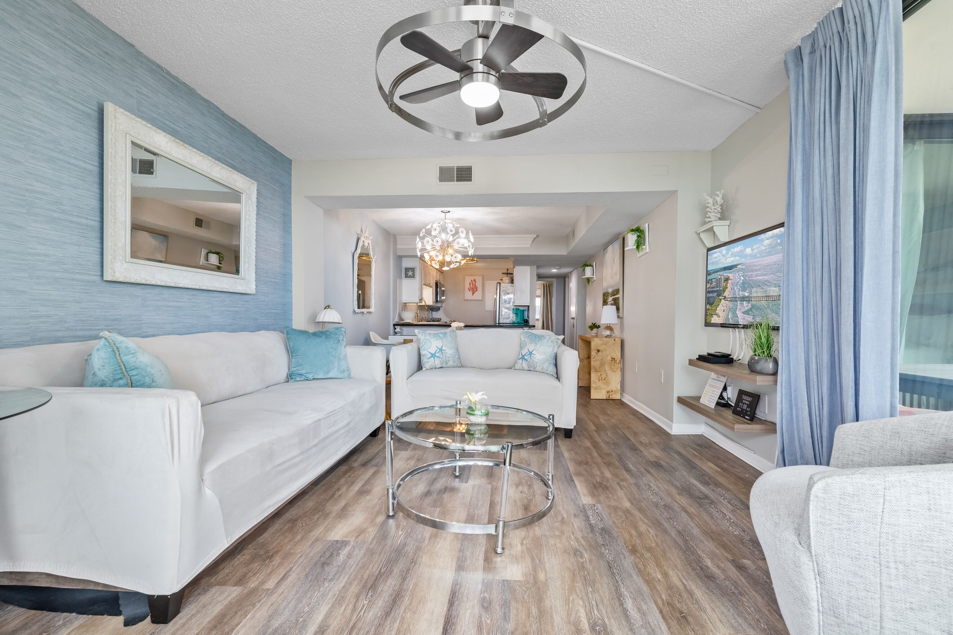 A modern living room with a white sofa, a glass coffee table, a ceiling fan, and a kitchen in the background.