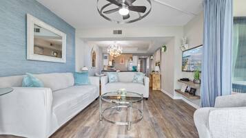 A modern living room with a white sofa, a glass coffee table, a ceiling fan, and a kitchen in the background.