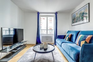 Un salon avec un téléviseur, un canapé bleu, une table basse et une fenêtre avec des rideaux.